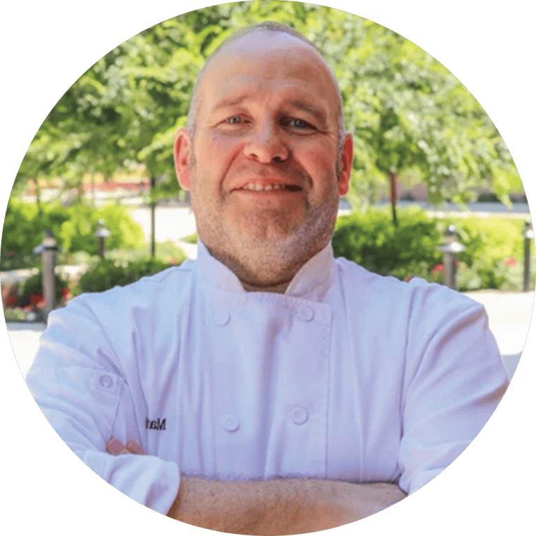 A chef in a white uniform stands outdoors with arms crossed, smiling, and greenery is visible in the background.
