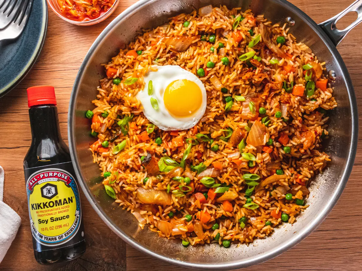 A skillet of fried rice with vegetables and a sunny-side-up egg on top sits next to a bottle of Kikkoman soy sauce.