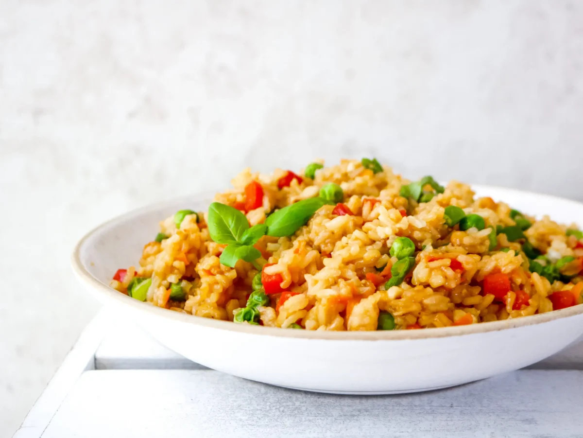 A white plate filled with vegetable fried rice, including peas and diced red bell peppers, garnished with fresh basil leaves.