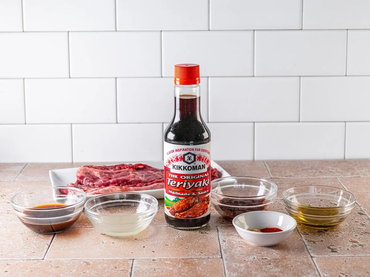 A bottle of Kikkoman Teriyaki marinade is placed on a counter, surrounded by raw ribs and small bowls containing various sauces and seasonings.