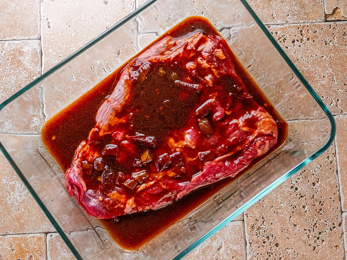 Raw steak marinating in a glass container with a dark, seasoned liquid on a tiled countertop.