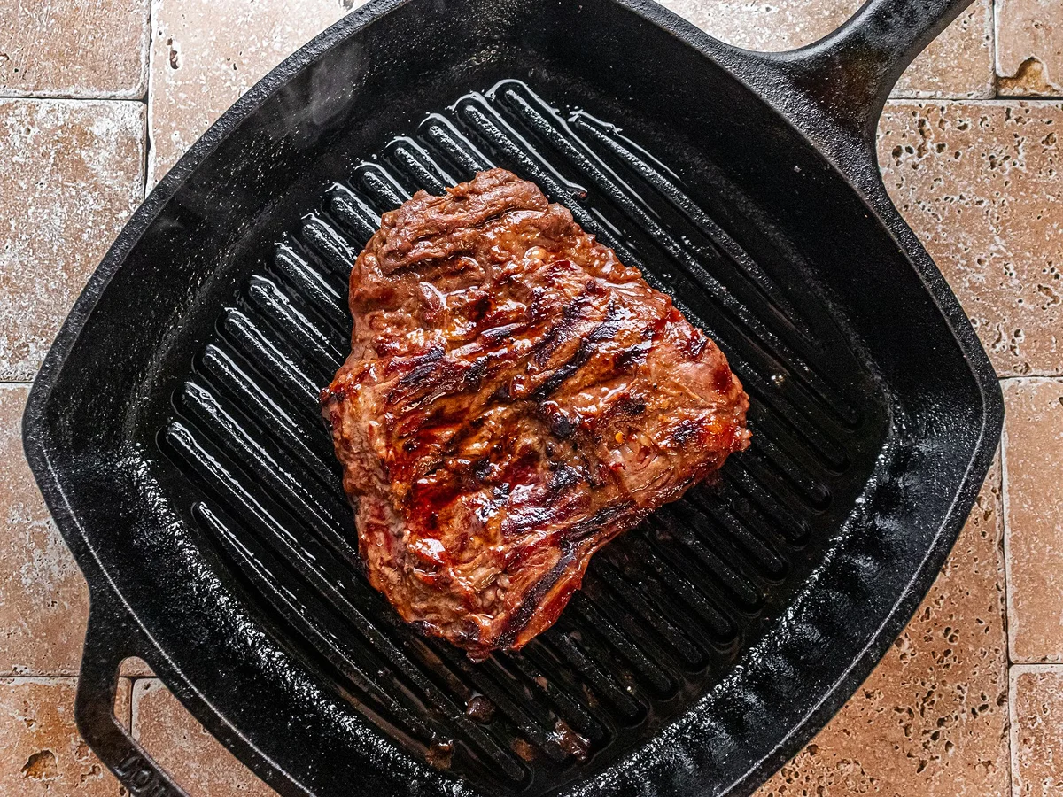 A grilled steak with dark sear marks sits in a black cast iron grill pan on a tiled surface.