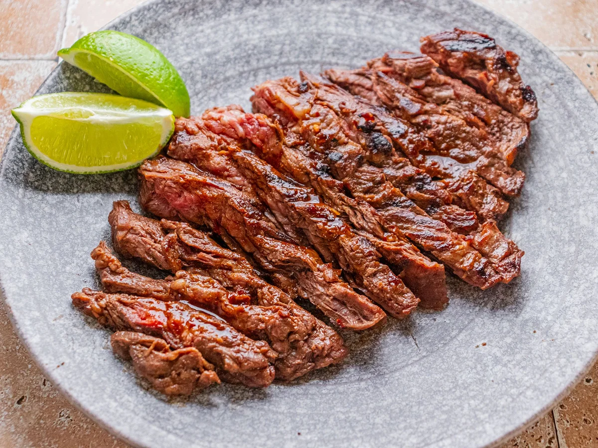 Sliced grilled steak served on a gray plate with two lime wedges.