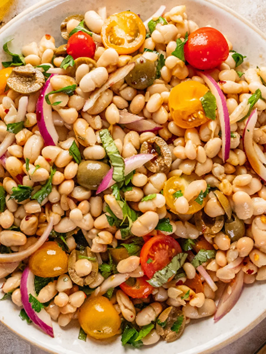 A bowl of white bean salad with cherry tomatoes, red onion, green olives, and fresh herbs.