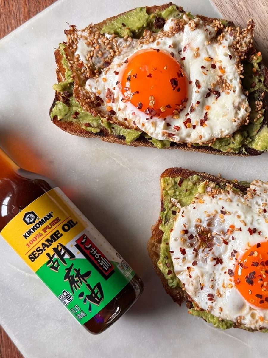 Two slices of avocado toast topped with sunny-side-up eggs, chili flakes, and sesame seeds, next to a bottle of Kikkoman sesame oil on a marble surface.