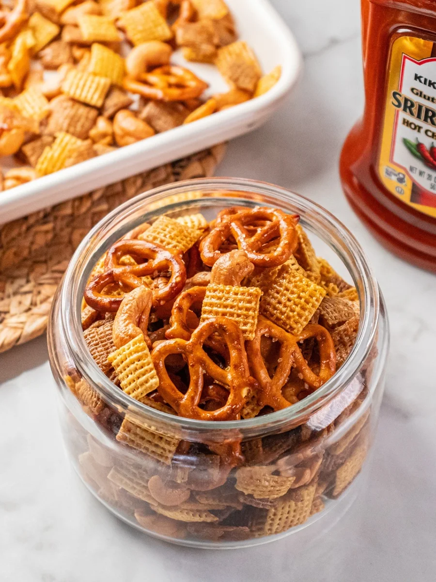 A glass jar filled with snack mix containing pretzels, Chex cereal, and nuts; a baking dish with more mix and a bottle of sriracha sauce are nearby.