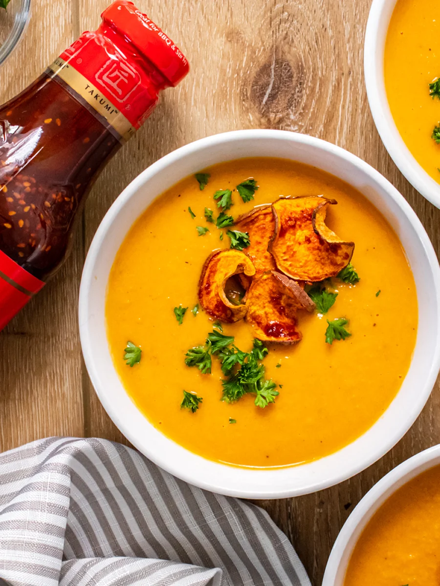 A bowl of orange soup topped with roasted squash slices and parsley sits on a wooden table next to a bottle of sauce and a striped napkin.