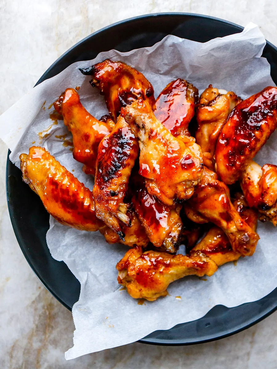 A black bowl lined with parchment paper holds a pile of glazed, roasted chicken wings on a light-colored surface.