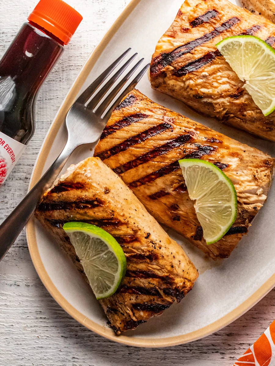 Three grilled salmon fillets with grill marks, each garnished with a lime wedge, served on an oval plate with a fork and knife; a bottle of sauce is nearby.