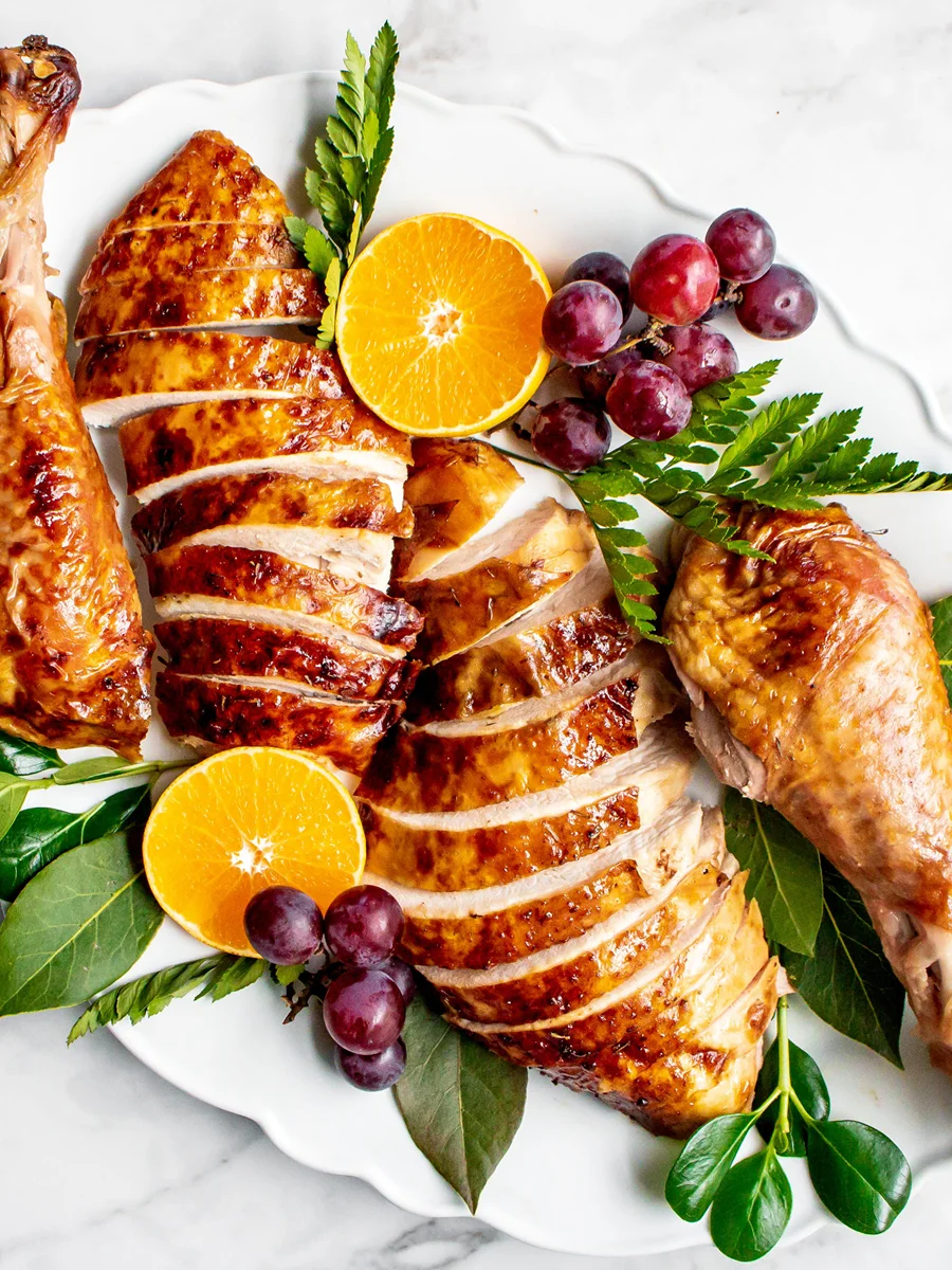 Sliced roasted turkey breast and drumsticks on a white platter, garnished with orange halves, grapes, and green leaves.