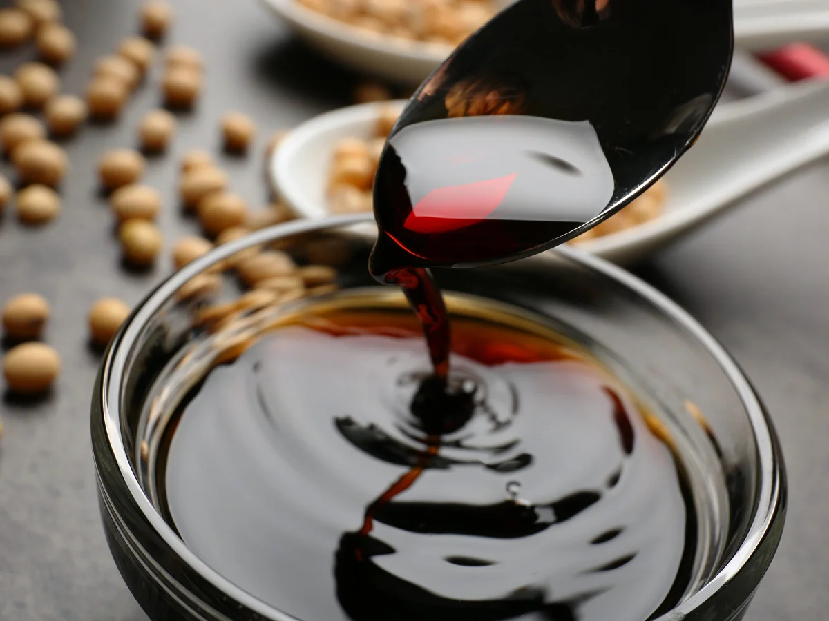 A spoon pours dark soy sauce into a glass bowl, with soybeans scattered and a white dish visible in the background.