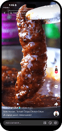 A hand dips a breaded chicken tender coated in sauce, shown in close-up on a smartphone screen displaying a food video.