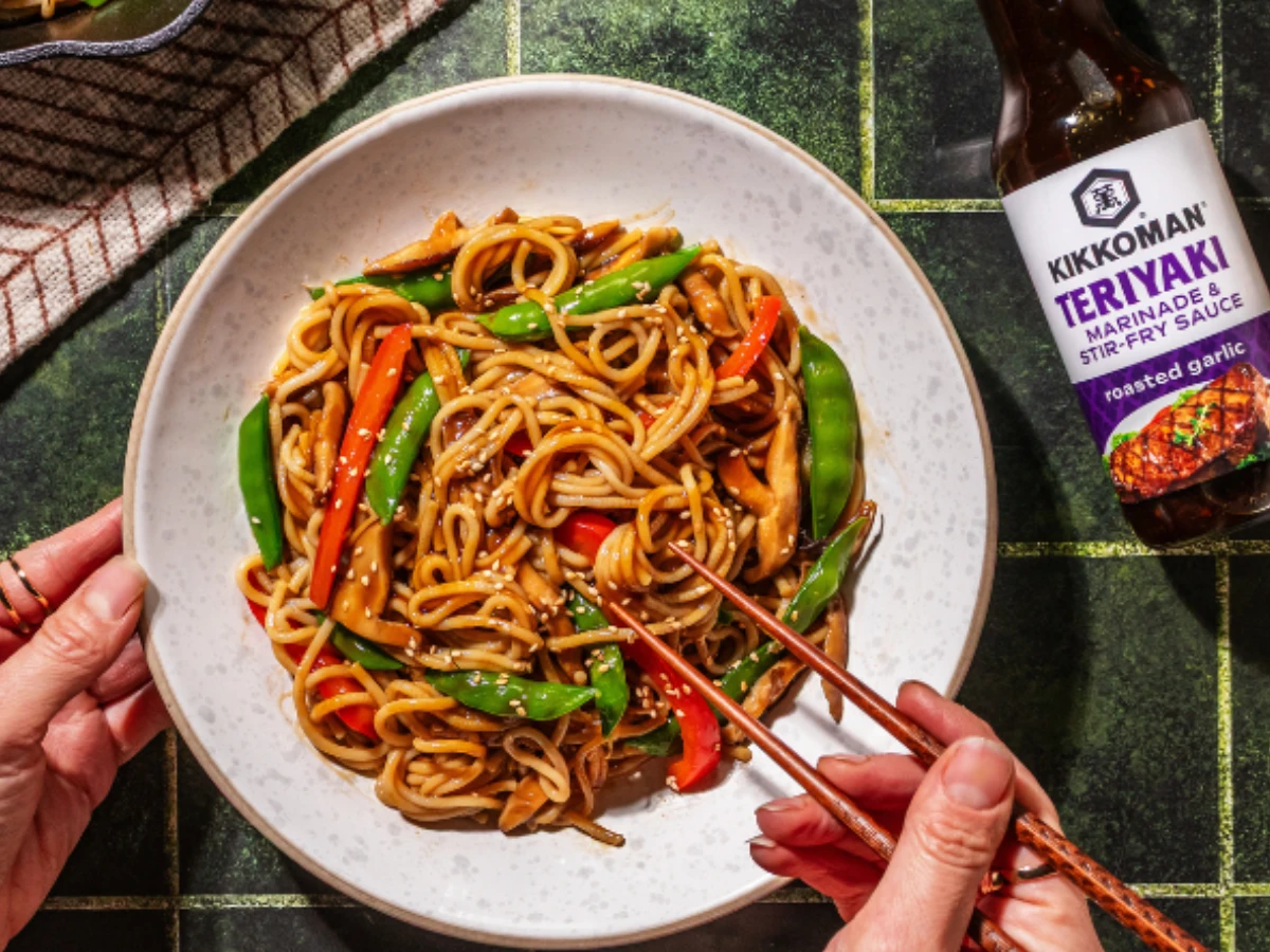 A person uses chopsticks to eat a plate of stir-fried noodles with vegetables. A bottle of Kikkoman teriyaki sauce is nearby on a green tiled surface.