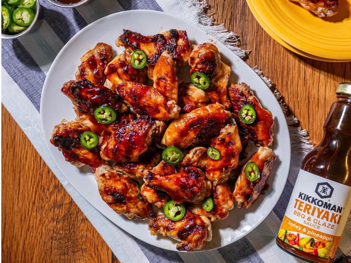 A platter of glazed chicken wings garnished with sliced jalapeños sits next to a bottle of Kikkoman Teriyaki BBQ & Glaze honey & pineapple sauce on a wooden table.