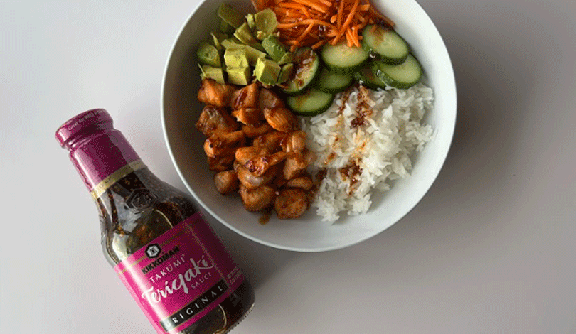 A bowl with white rice, grilled chicken, sliced cucumber, shredded carrots, and diced avocado next to a bottle of Takumi Teriyaki Sauce.