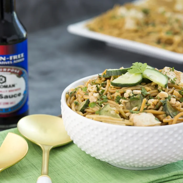 A white bowl filled with noodle stir-fry, garnished with cucumber slices and cilantro, sits next to a gold spoon and a bottle of soy sauce.