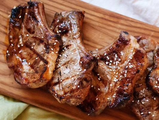 Grilled pork chops with grill marks, topped with sesame seeds, arranged on a wooden serving board.