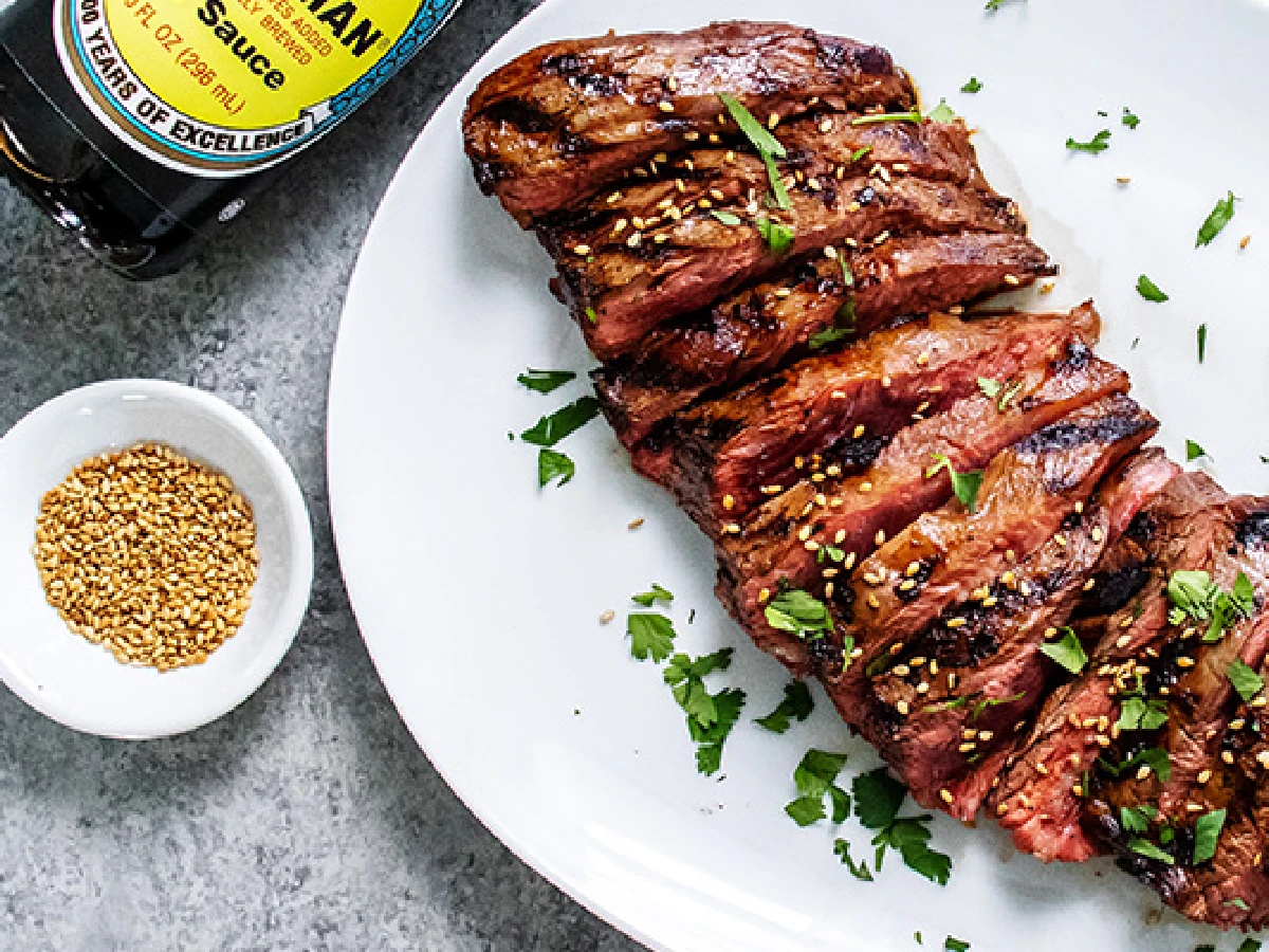 Sliced grilled steak garnished with sesame seeds and chopped herbs on a white plate, with a small bowl of sesame seeds and a bottle of soy sauce nearby.