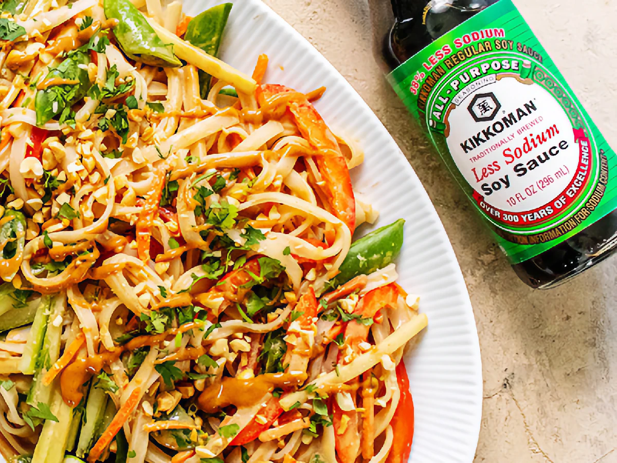 Plate of noodle salad with vegetables and peanuts, next to a bottle of Kikkoman Less Sodium Soy Sauce on a white surface.