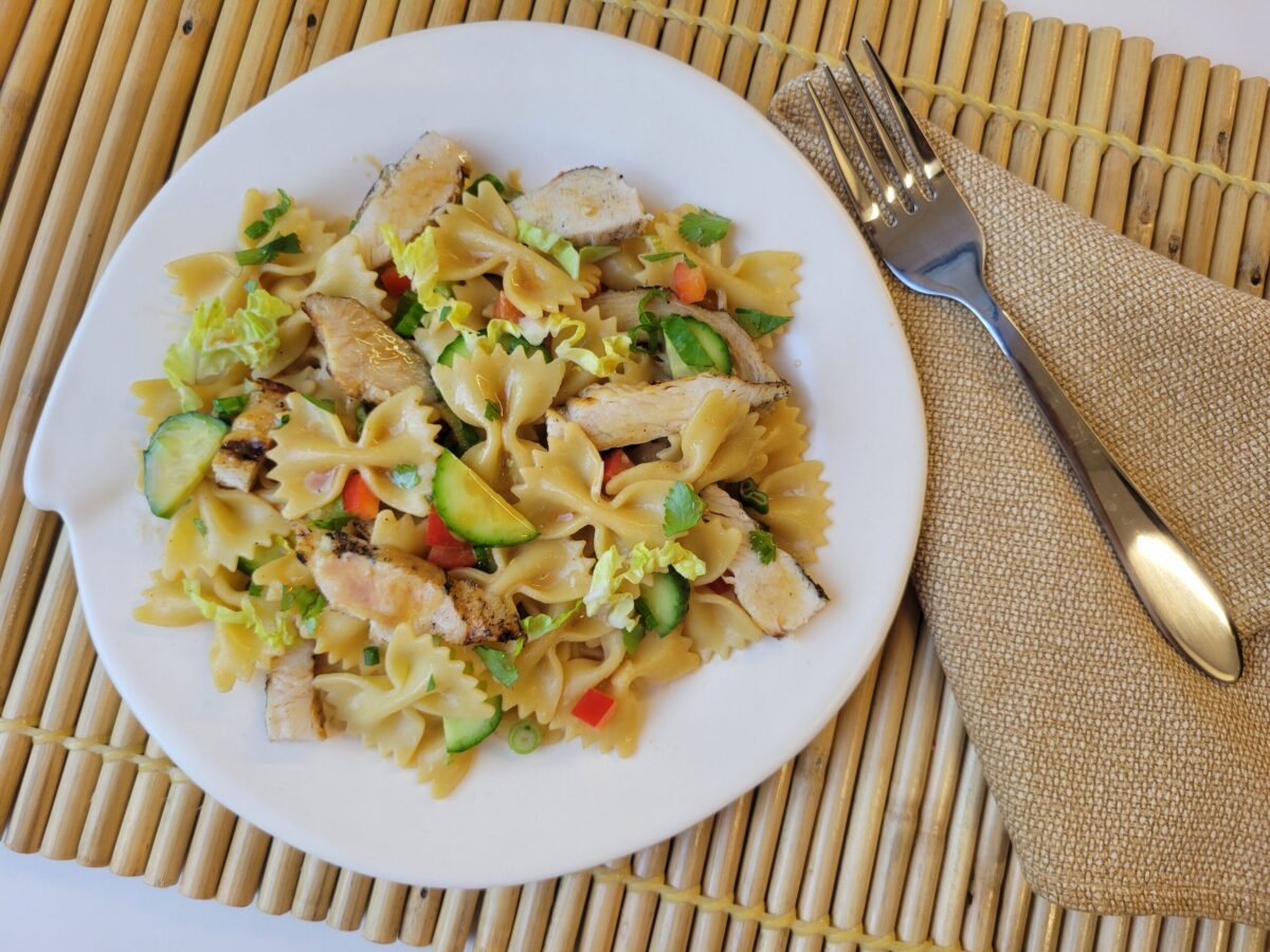 A white plate with bowtie pasta, grilled chicken strips, sliced zucchini, carrots, and green onions, placed on a bamboo mat with a fork and napkin beside it.