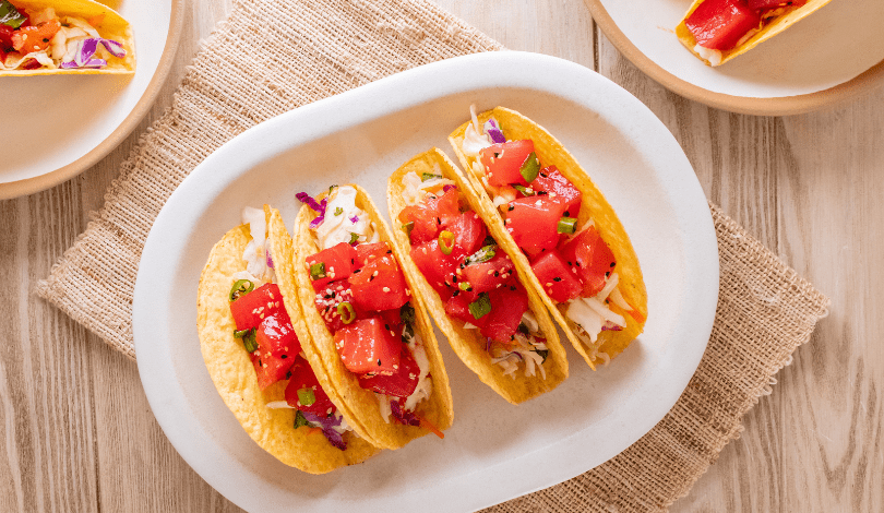 Three hard shell tacos filled with shredded cabbage, diced tomatoes, and green onions are arranged on a white plate atop a woven placemat.
