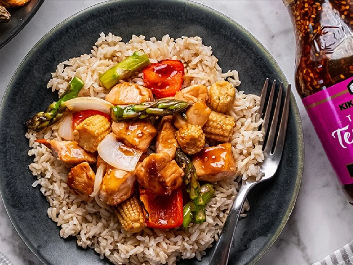A plate of brown rice topped with stir-fried chicken, baby corn, asparagus, onions, and red bell peppers, with a fork and a bottle of teriyaki sauce nearby.