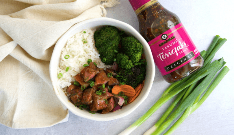 A bowl of white rice, broccoli, carrots, and teriyaki beef is placed next to a bottle of teriyaki sauce, green onions, and a beige napkin on a light surface.