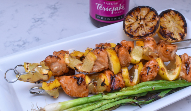 Grilled chicken and yellow pepper skewers served with charred lemon halves and green onions on a white plate; teriyaki sauce bottle in the background.