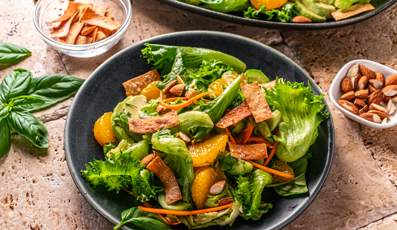 A bowl of green salad with mandarin oranges, sliced carrots, tortilla strips, and chopped almonds, with basil leaves and small bowls of tortilla strips and almonds on the side.
