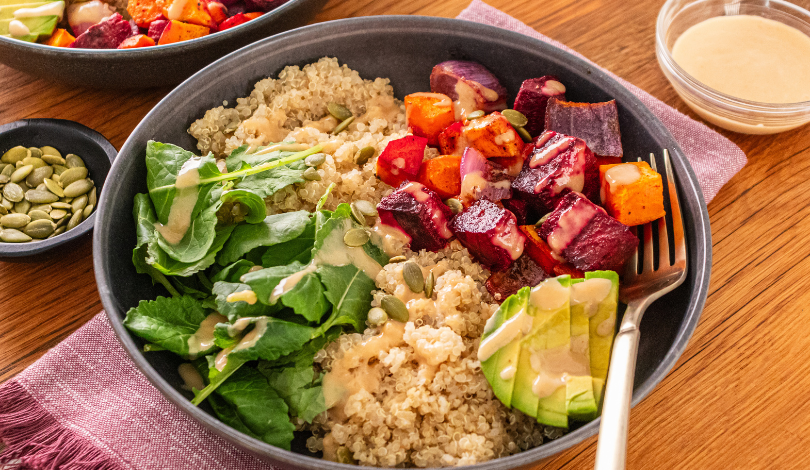 A bowl with quinoa, fresh greens, sliced avocado, roasted root vegetables, pumpkin seeds, and a drizzle of dressing, served with a fork on a wooden table.