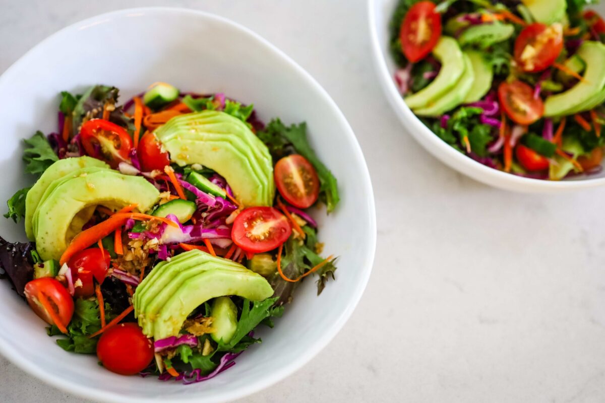Two white bowls filled with salad containing avocado slices, cherry tomatoes, shredded carrots, cabbage, and mixed greens on a light surface.