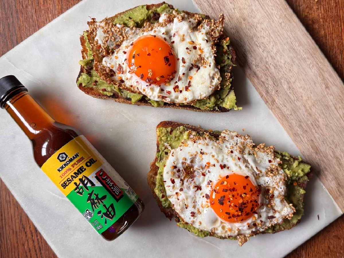 Two slices of toast topped with mashed avocado, sunny-side-up eggs, and red pepper flakes sit on parchment paper next to a bottle of Kikkoman sesame oil.