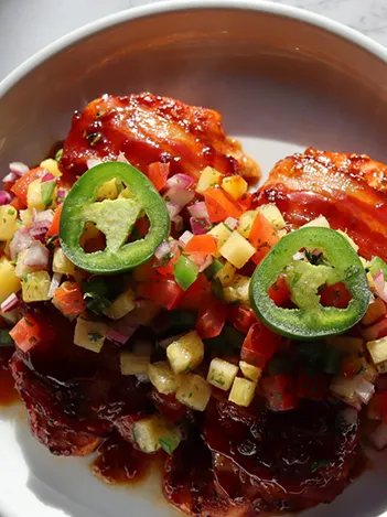 Barbecued chicken thighs topped with a colorful diced vegetable and fruit salsa, garnished with fresh jalapeño slices, served on a white plate.