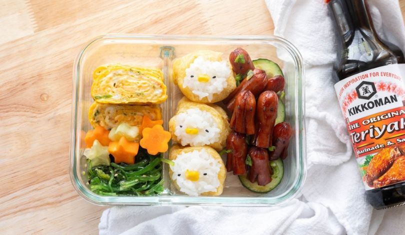 A glass bento box with rolled omelet, rice balls shaped like chicks, mini sausages, sliced cucumber, spinach, and a carrot flower, next to a bottle of teriyaki sauce.