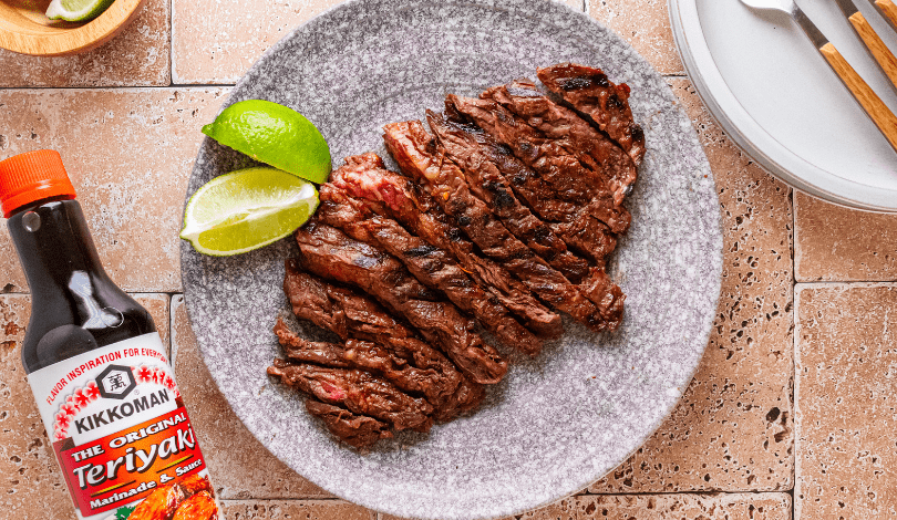 Sliced grilled steak served with two lime wedges on a gray plate, next to a bottle of Kikkoman Teriyaki Marinade & Sauce.