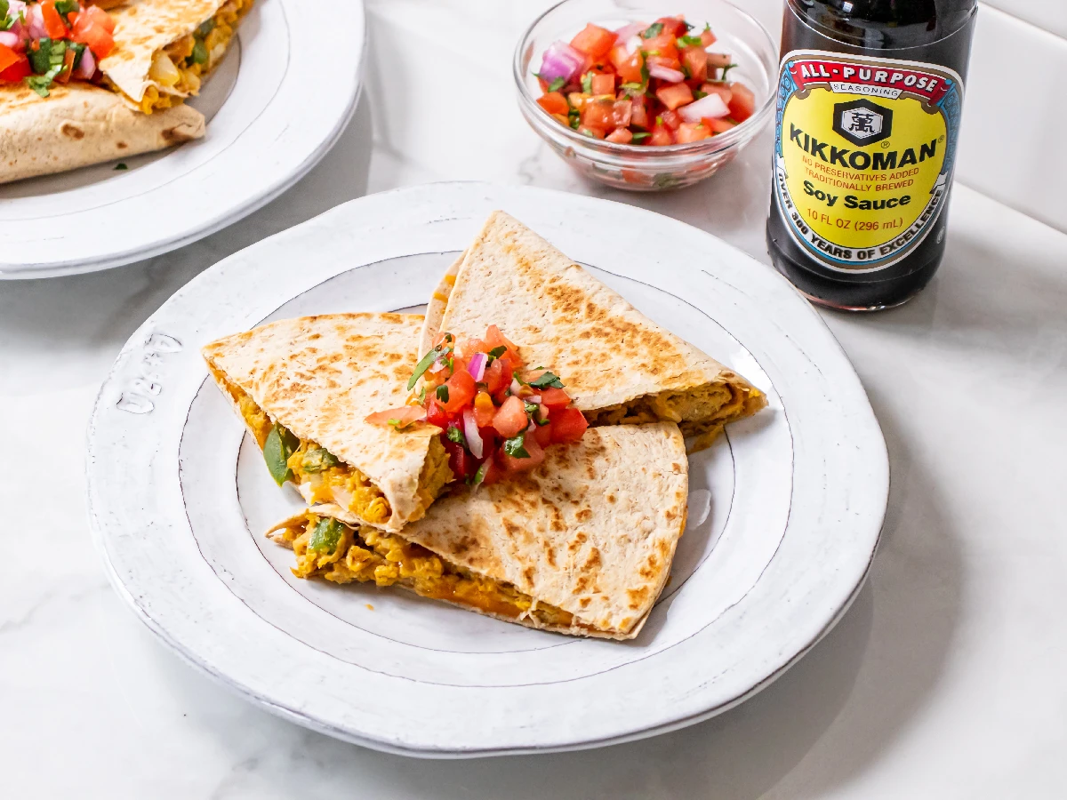 A plate with quesadilla wedges topped with pico de gallo, next to a bowl of salsa and a bottle of Kikkoman soy sauce on a white surface.