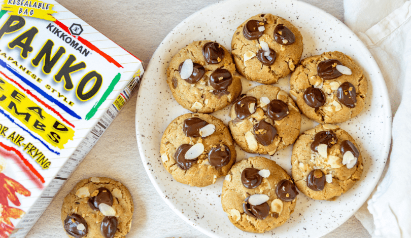 A plate of cookies topped with chocolate and almond slices sits next to a box of Kikkoman Panko bread crumbs; one cookie is placed beside the plate.