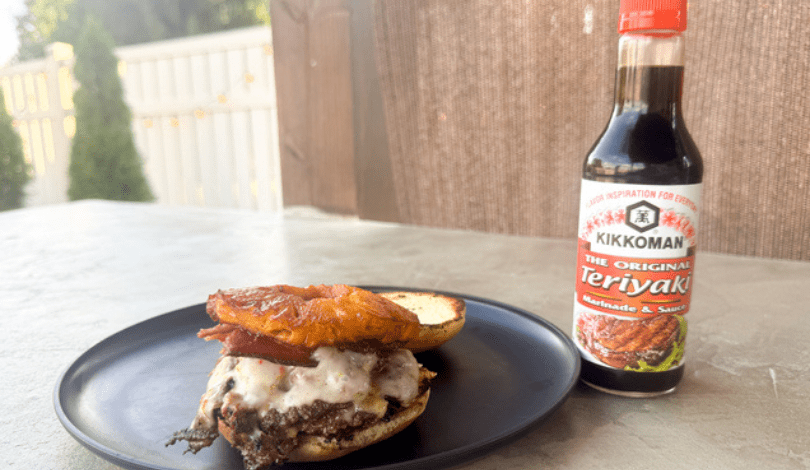 A cheeseburger with bacon and pineapple sits on a black plate next to a bottle of Kikkoman Teriyaki sauce on an outdoor table.