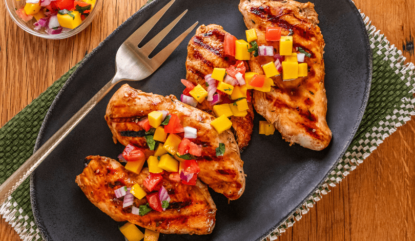 Three grilled chicken breasts topped with diced mango salsa are arranged on a black plate with a large fork, placed on a woven green and white placemat.