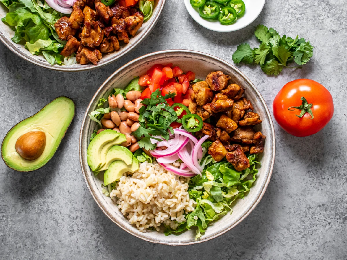 A bowl with grilled chicken, brown rice, lettuce, avocado, pinto beans, diced tomatoes, pickled onions, and cilantro, with fresh toppings like tomato, jalapeño, and cilantro beside it.