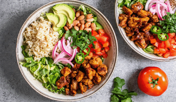 30 Minutes or Less Two bowls with rice, grilled chicken, avocado, pinto beans, lettuce, tomatoes, pickled onions, cilantro, and jalapeño slices. Tomato and cilantro garnish on the side.