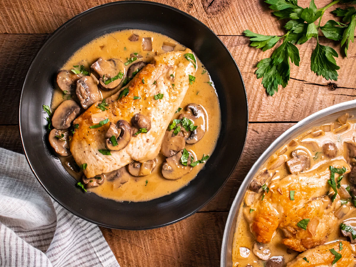 Chicken breast in a creamy mushroom sauce garnished with parsley, served on a black plate, with a striped napkin and fresh parsley on the wooden table.
