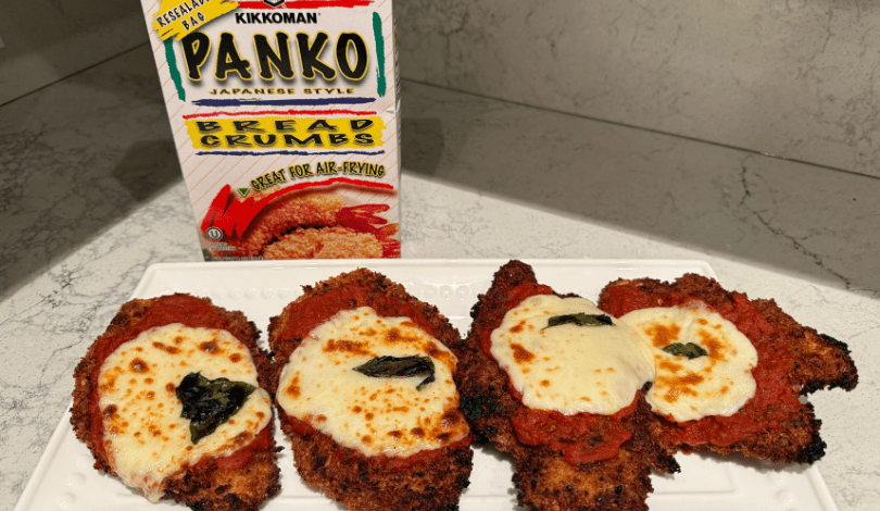 Four pieces of chicken parmesan with melted cheese and basil are arranged on a white plate. A box of Kikkoman panko bread crumbs is in the background.