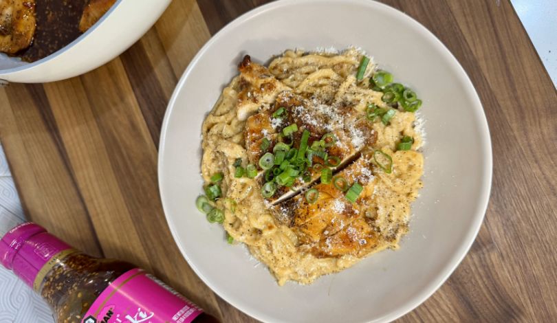 A bowl of creamy pasta topped with grilled chicken, chopped green onions, and grated cheese sits on a wooden surface next to a bottle of sauce.