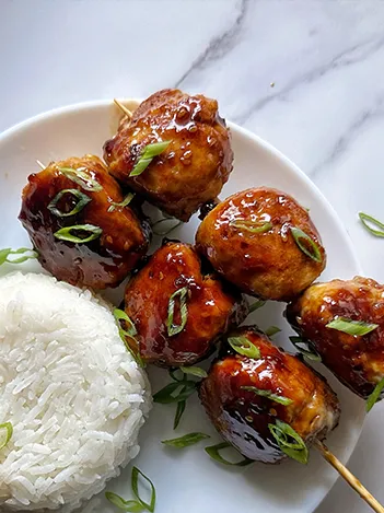 A plate with two skewers of glazed meatballs garnished with sliced green onions, served next to a mound of white rice on a white surface.