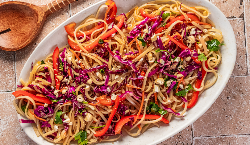 A white oval dish with noodles, sliced red bell peppers, shredded purple cabbage, chopped herbs, and crushed peanuts, set on a tiled surface next to a wooden spoon.