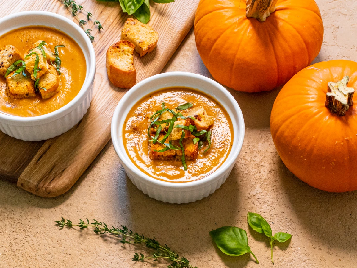 Two bowls of creamy pumpkin soup topped with croutons and herbs, placed beside fresh pumpkins and basil leaves on a beige surface.
