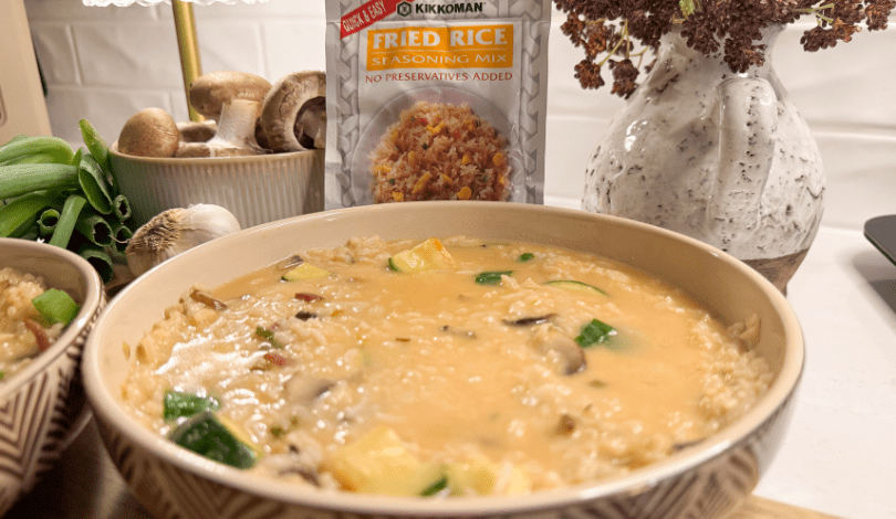 A bowl of creamy rice with vegetables sits in front of a Kikkoman Fried Rice Seasoning Mix packet, surrounded by fresh mushrooms and green onions on a kitchen counter.