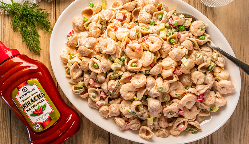 A plate of creamy pasta salad with chopped vegetables sits next to a bottle of sriracha-style hot sauce on a wooden table.