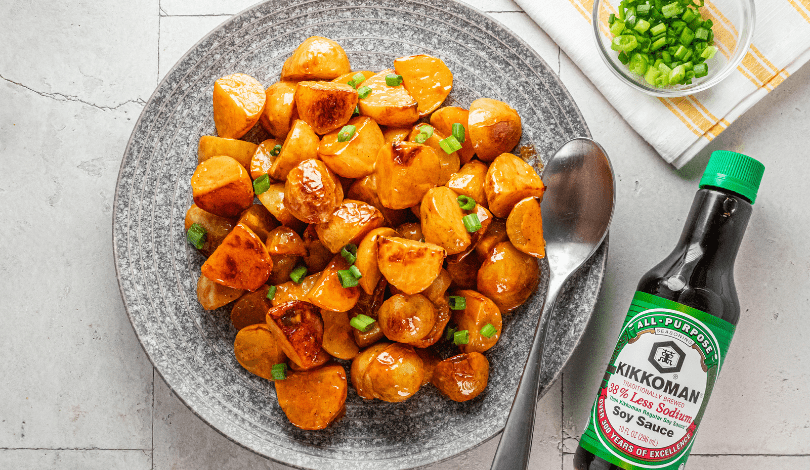 A plate of glazed roasted potatoes garnished with chopped green onions, a spoon, a bottle of soy sauce, and a small bowl of green onions on the side.